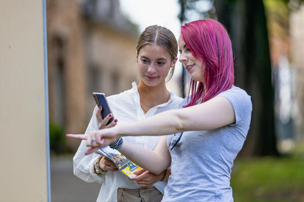 Bild vergrößern: Zwei junge Frauen stehen mit dem Handy vor der Infotafel am Startpunkt des Rundwegs "Zwischen den Mauern". Im Hintergrund ist das Korbacher Museum zu sehen.