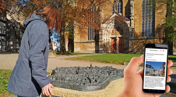 Bild vergrößern: Eine junge Frau steht am Bronze-Stadtmodell auf dem Kirchplatz Korbach. Neben ihr ist ein Handy mit der Startseite der Audioguide-Stadtführung durch Korbach eingeblendet.