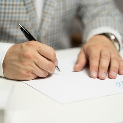 Bild vergrößern: Symbolbild Ausschreibungen
Ein Mann in einem grau karrierten Anzug unterschreibt einen Zettel. In der Mitte des Bildes sind seine Hände abgebildet. In der linken Hand hält er einen schwarzen Kugelschreiber.